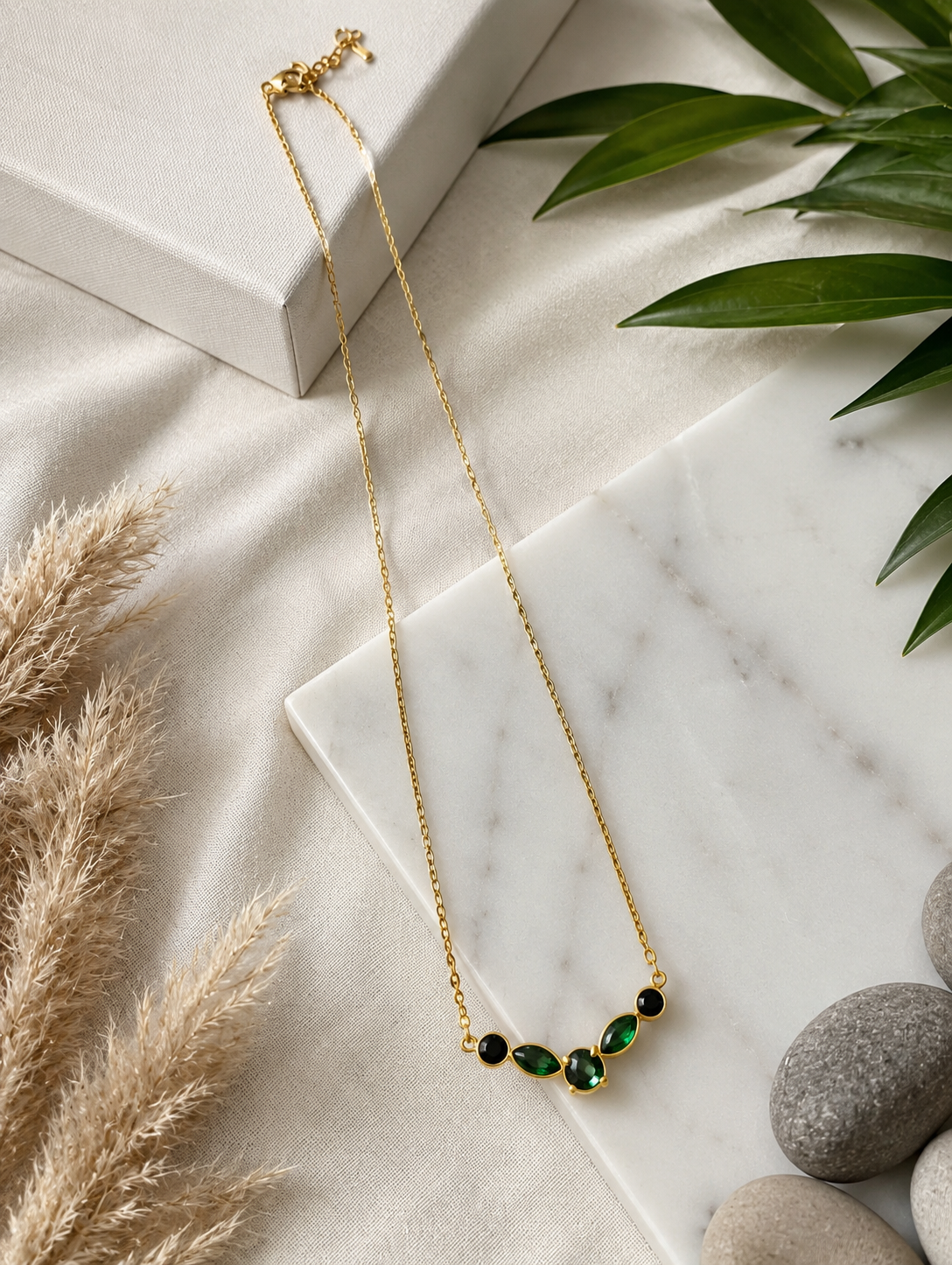 A product photography collage featuring a delicate gold-toned necklace with a curved station pendant of five faceted gemstones in rich emerald green and deep black. The first view shows a close-up of the necklace resting on a white marble slab with smooth stones and textured linen. The second shows the entire length of the gold link chain in a styled flat-lay next to a gift box and tropical leaves. The third shows the necklace being worn by a model, resting elegantly on her collarbone with a taupe off-the-s