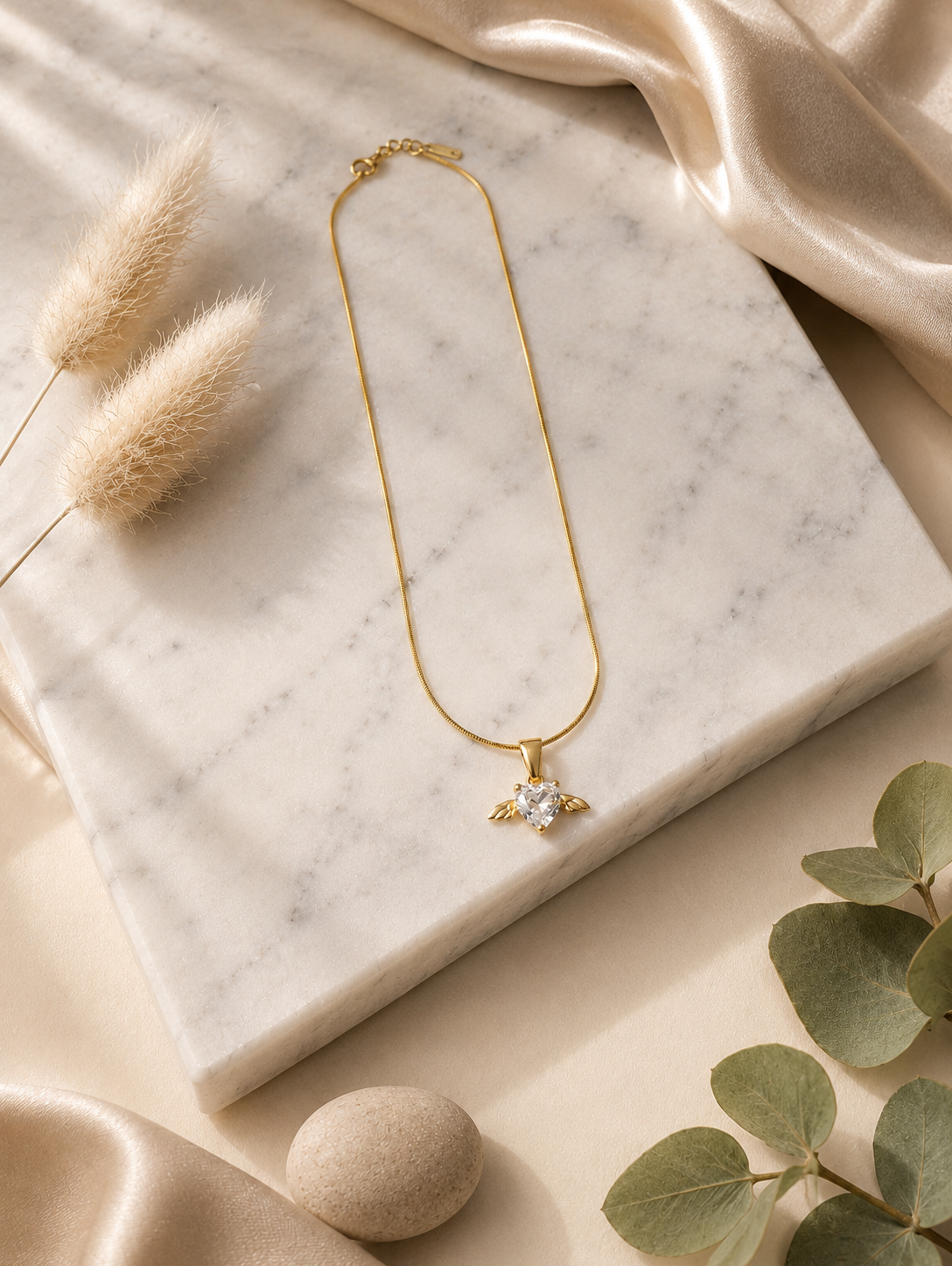 A product photography display showcasing a delicate gold-toned snake chain necklace with an angel wing heart pendant. The collection includes a styled flat-lay of the necklace on a white marble slab surrounded by champagne silk and dried botanical elements, a macro close-up highlighting the faceted clear crystal heart and its gold wings, and a lifestyle image of a model wearing the necklace on her collarbone.