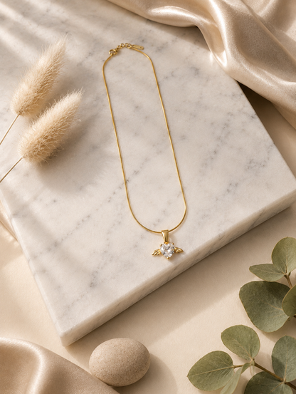 A product photography display showcasing a delicate gold-toned snake chain necklace with an angel wing heart pendant. The collection includes a styled flat-lay of the necklace on a white marble slab surrounded by champagne silk and dried botanical elements, a macro close-up highlighting the faceted clear crystal heart and its gold wings, and a lifestyle image of a model wearing the necklace on her collarbone.