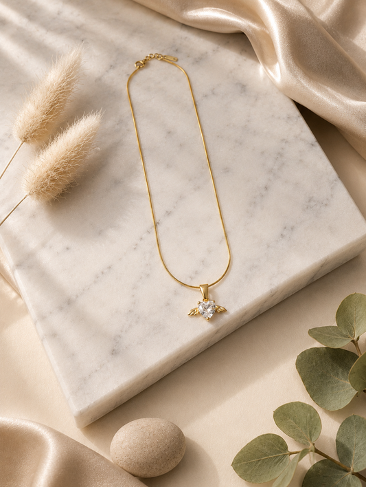 A product photography display showcasing a delicate gold-toned snake chain necklace with an angel wing heart pendant. The collection includes a styled flat-lay of the necklace on a white marble slab surrounded by champagne silk and dried botanical elements, a macro close-up highlighting the faceted clear crystal heart and its gold wings, and a lifestyle image of a model wearing the necklace on her collarbone.