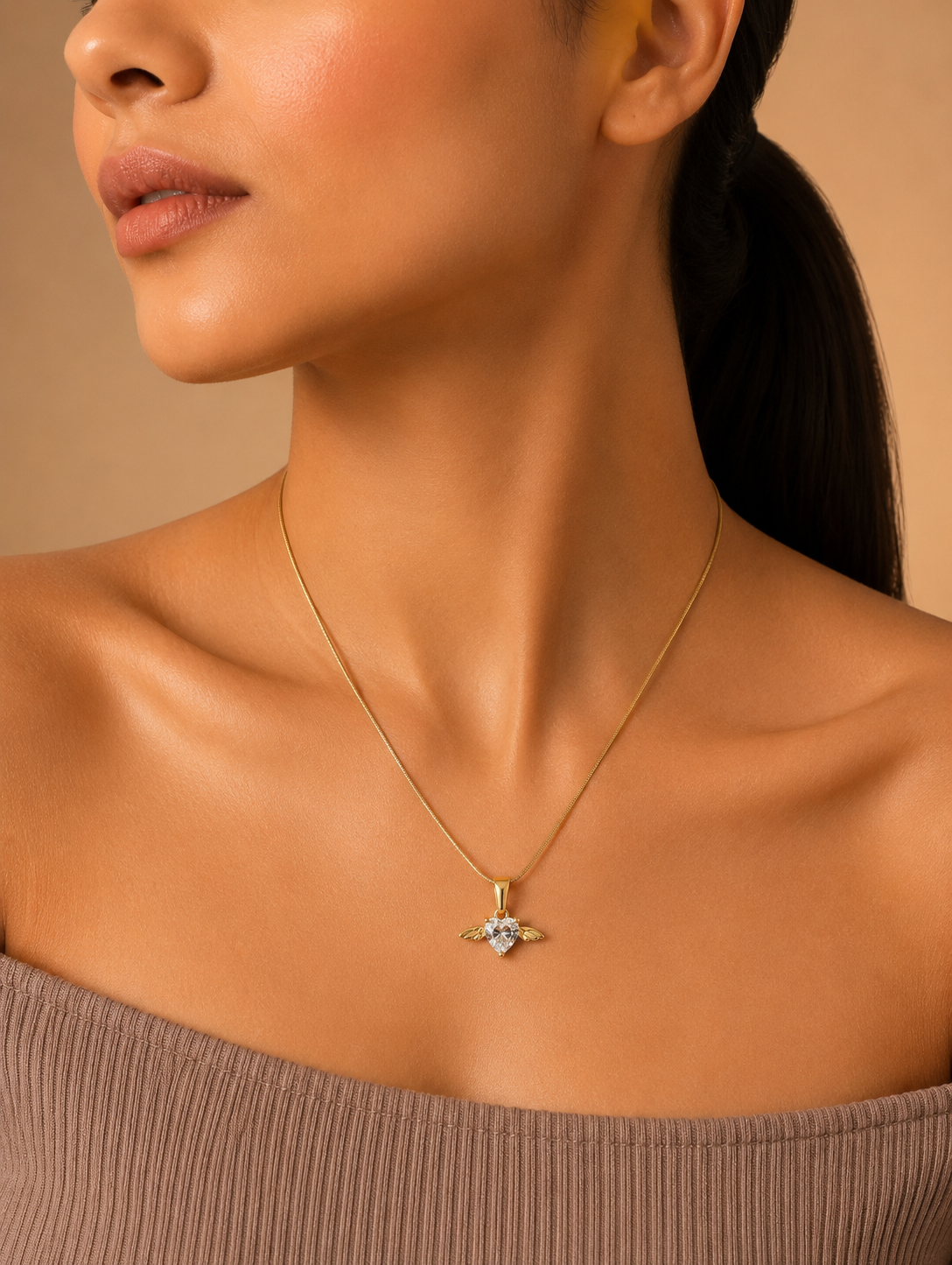 A product photography display showcasing a delicate gold-toned snake chain necklace with an angel wing heart pendant. The collection includes a styled flat-lay of the necklace on a white marble slab surrounded by champagne silk and dried botanical elements, a macro close-up highlighting the faceted clear crystal heart and its gold wings, and a lifestyle image of a model wearing the necklace on her collarbone.