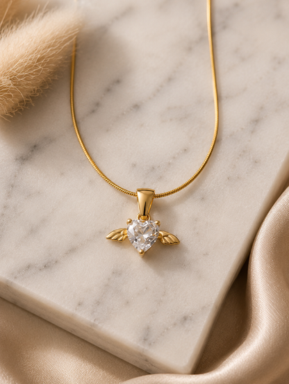 A product photography display showcasing a delicate gold-toned snake chain necklace with an angel wing heart pendant. The collection includes a styled flat-lay of the necklace on a white marble slab surrounded by champagne silk and dried botanical elements, a macro close-up highlighting the faceted clear crystal heart and its gold wings, and a lifestyle image of a model wearing the necklace on her collarbone.