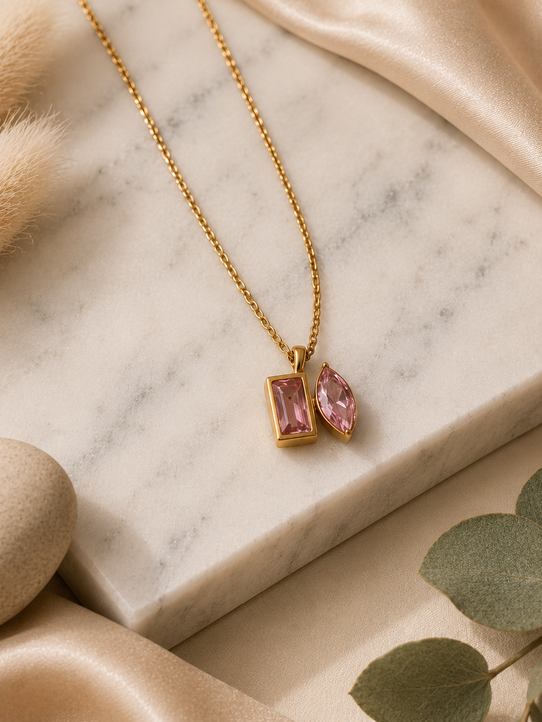 A product photography showcase of a gold-toned link chain necklace featuring a unique dual-cut pink crystal pendant with one rectangular and one marquise-cut stone. The images include a close-up detail shot on a marble surface, a full flat-lay arrangement with dried botanicals and eucalyptus, and a display view highlighting how the necklace hangs on a cream-colored jewelry bust.