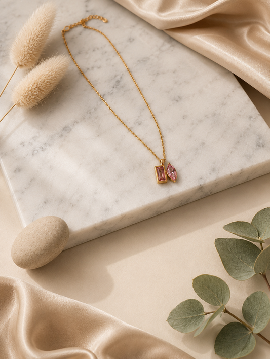 A product photography showcase of a gold-toned link chain necklace featuring a unique dual-cut pink crystal pendant with one rectangular and one marquise-cut stone. The images include a close-up detail shot on a marble surface, a full flat-lay arrangement with dried botanicals and eucalyptus, and a display view highlighting how the necklace hangs on a cream-colored jewelry bust.