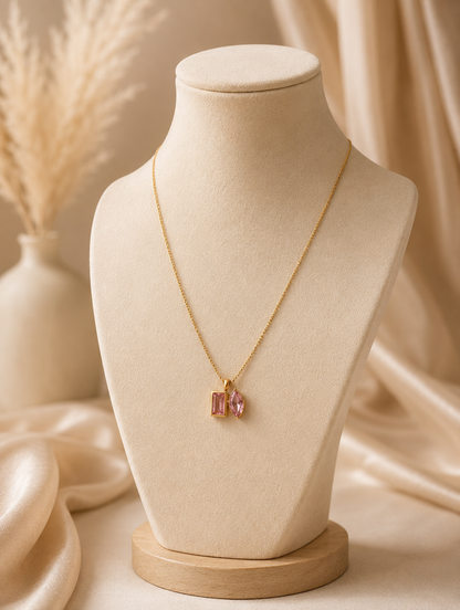 A product photography showcase of a gold-toned link chain necklace featuring a unique dual-cut pink crystal pendant with one rectangular and one marquise-cut stone. The images include a close-up detail shot on a marble surface, a full flat-lay arrangement with dried botanicals and eucalyptus, and a display view highlighting how the necklace hangs on a cream-colored jewelry bust.