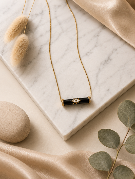 A product photography display showcasing a modern gold-toned necklace with a horizontal black enamel bar evil eye pendant. The collection includes a styled flat-lay view on a marble slab surrounded by champagne satin and dried botanicals, a display view of the necklace resting on a cream velvet jewelry mannequin, and a macro close-up highlighting the crisp white, black, and gold details of the central eye motif and its textured end caps.