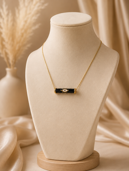 A product photography display showcasing a modern gold-toned necklace with a horizontal black enamel bar evil eye pendant. The collection includes a styled flat-lay view on a marble slab surrounded by champagne satin and dried botanicals, a display view of the necklace resting on a cream velvet jewelry mannequin, and a macro close-up highlighting the crisp white, black, and gold details of the central eye motif and its textured end caps.