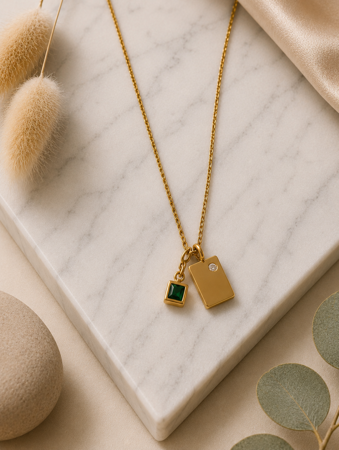 A product photography showcase of a delicate gold-toned chain necklace featuring a dual geometric charm pendant—a smooth rectangular gold tag with a tiny inset crystal next to a princess-cut emerald green square charm. The collection includes a close-up detail view on a marble slab, a styled lifestyle flat-lay with dried botanical accents, and a model display view showing how the necklace rests gracefully on the collarbone.