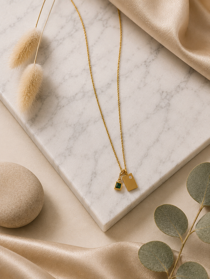 A product photography showcase of a delicate gold-toned chain necklace featuring a dual geometric charm pendant—a smooth rectangular gold tag with a tiny inset crystal next to a princess-cut emerald green square charm. The collection includes a close-up detail view on a marble slab, a styled lifestyle flat-lay with dried botanical accents, and a model display view showing how the necklace rests gracefully on the collarbone.