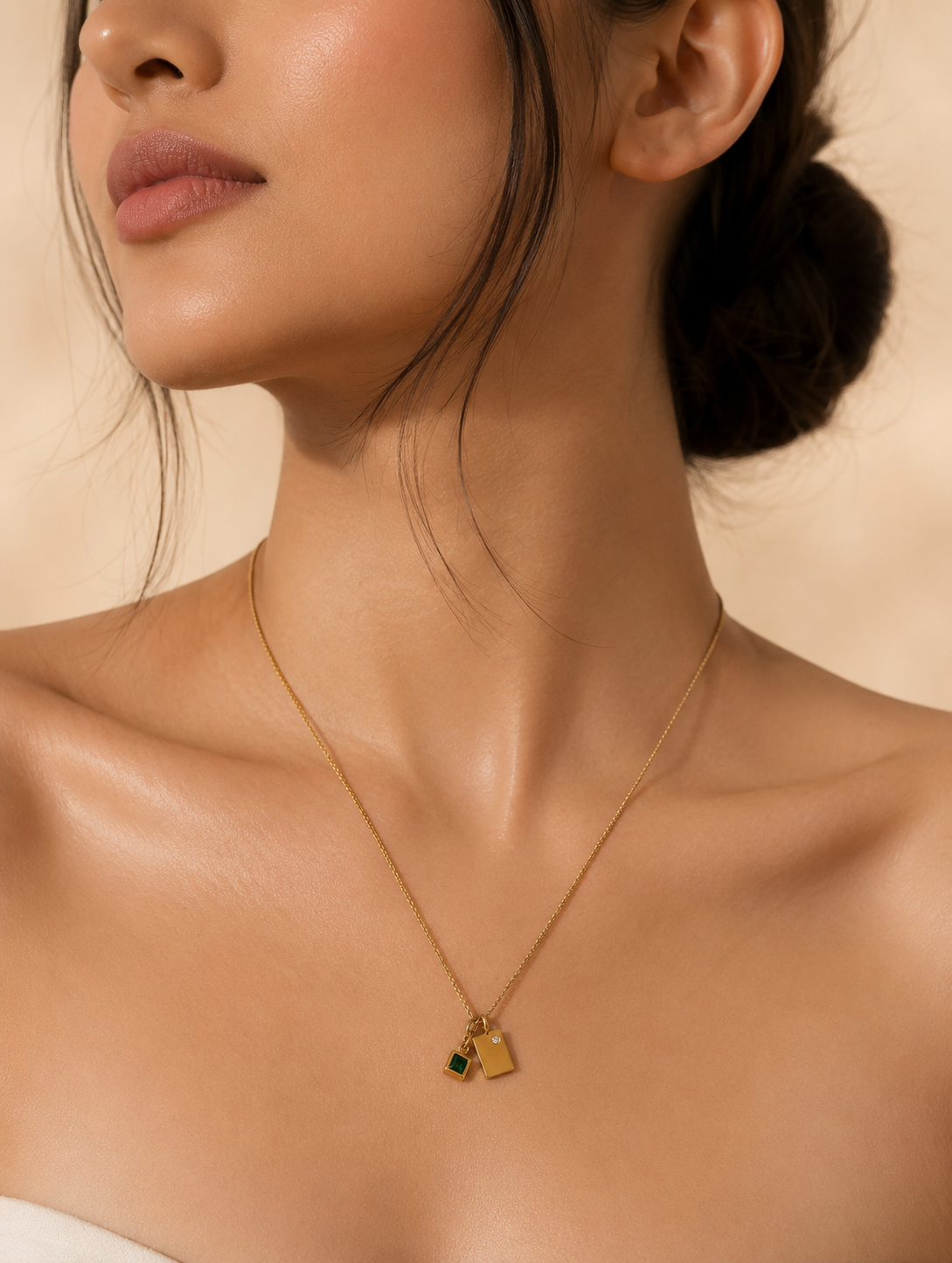 A product photography showcase of a delicate gold-toned chain necklace featuring a dual geometric charm pendant—a smooth rectangular gold tag with a tiny inset crystal next to a princess-cut emerald green square charm. The collection includes a close-up detail view on a marble slab, a styled lifestyle flat-lay with dried botanical accents, and a model display view showing how the necklace rests gracefully on the collarbone.