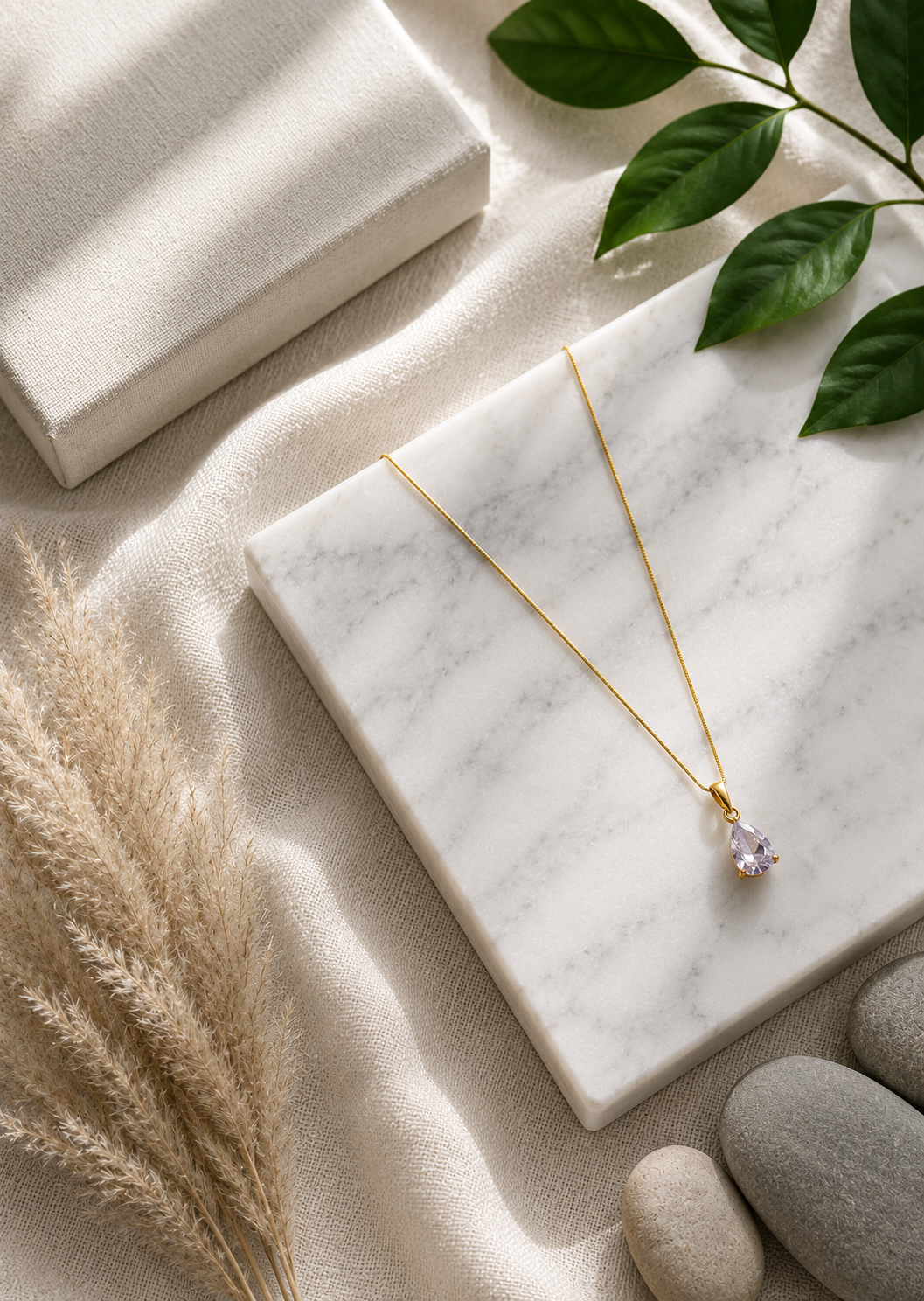 A product photography showcase of a delicate gold-toned necklace featuring a faceted lavender teardrop crystal pendant. The collection includes a styled flat-lay view on a marble block with dried botanicals and river pebbles, a display view of the necklace on a black velvet jewelry bust against champagne silk, and a macro close-up detailing the pear-cut crystal facets and its polished gold-toned prong setting.
