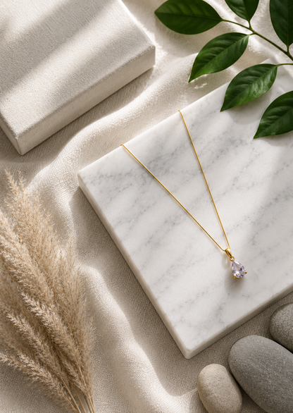 A product photography showcase of a delicate gold-toned necklace featuring a faceted lavender teardrop crystal pendant. The collection includes a styled flat-lay view on a marble block with dried botanicals and river pebbles, a display view of the necklace on a black velvet jewelry bust against champagne silk, and a macro close-up detailing the pear-cut crystal facets and its polished gold-toned prong setting.