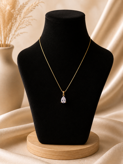 A product photography showcase of a delicate gold-toned necklace featuring a faceted lavender teardrop crystal pendant. The collection includes a styled flat-lay view on a marble block with dried botanicals and river pebbles, a display view of the necklace on a black velvet jewelry bust against champagne silk, and a macro close-up detailing the pear-cut crystal facets and its polished gold-toned prong setting.