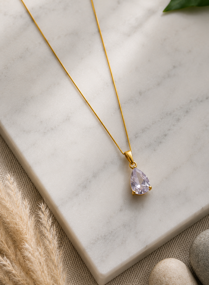 A product photography showcase of a delicate gold-toned necklace featuring a faceted lavender teardrop crystal pendant. The collection includes a styled flat-lay view on a marble block with dried botanicals and river pebbles, a display view of the necklace on a black velvet jewelry bust against champagne silk, and a macro close-up detailing the pear-cut crystal facets and its polished gold-toned prong setting.