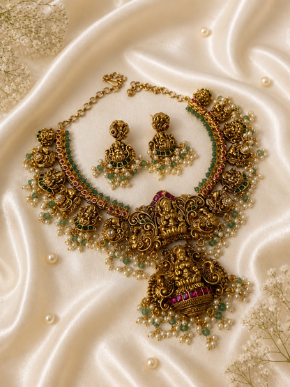 Antique matte finish gold necklace and earring set featuring intricate carvings of Goddess Lakshmi, accented with emerald green and ruby pink beads and delicate hanging pearl clusters, displayed on a draped cream satin background.