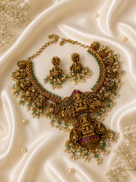 Antique matte finish gold necklace and earring set featuring intricate carvings of Goddess Lakshmi, accented with emerald green and ruby pink beads and delicate hanging pearl clusters, displayed on a draped cream satin background.