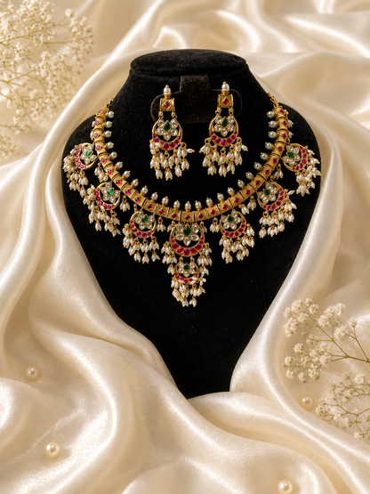 A royal Maharani style gold choker necklace held in a hand, showcasing crescent-shaped Jadau Kundan pendants set with ruby pink and emerald green stones, finished with dense clusters of small white pearls.
