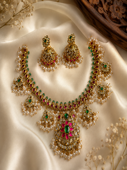 Madura traditional Indian jewelry set displayed on a black bust mannequin, featuring an antique gold necklace with a vibrant pink and green gemstone floral pendant, tiered pearl drops, and matching dome-shaped jhumka earrings.