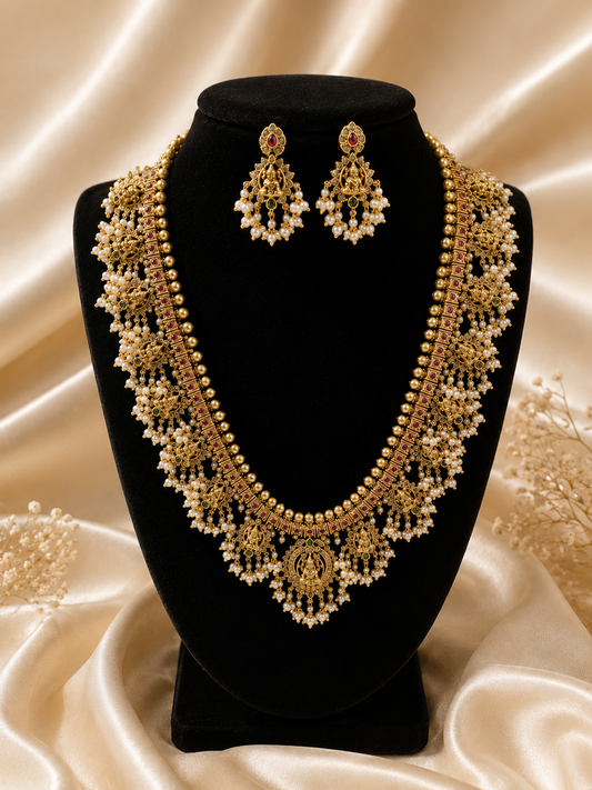 Exquisite long South Indian temple jewelry haram necklace and earring set on a black display stand, featuring rows of golden beads, intricate deity engravings, kemp stone detailing, and elaborate pearl cluster extensions.