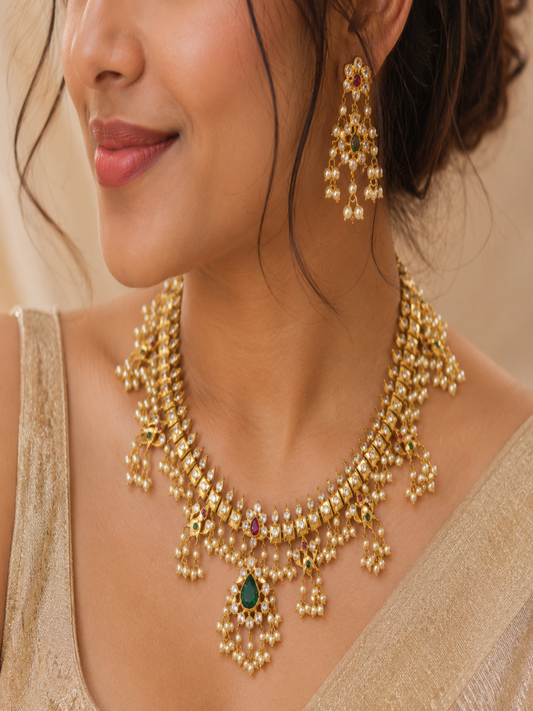 Close-up of a model wearing the Ramba short traditional Indian gold necklace and matching chandelier earrings, beautifully adorned with square Kundan stone settings, a central green enamel drop, and dangling clusters of delicate white pearls.