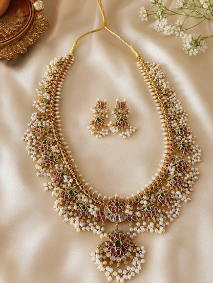 Flat lay view of the Revathy Long Guttapusalu necklace and its matching earrings laid gracefully on a shimmering cream fabric backdrop next to decorative gold plates Flat lay view of the Revathy Long Guttapusalu necklace and its matching earrings laid gracefully on a shimmering cream fabric backdrop next to decorative gold plates and white flowers.and white flowers.