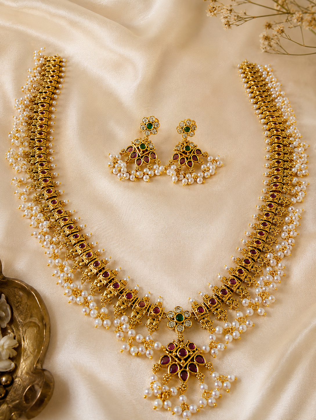 A flat lay view of the Manushya Guttapusalu long necklace and matching floral-infused drop earrings arranged gracefully on shimmering cream silk fabric, emphasizing the ruby gemstone detailing.