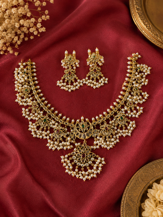 A flat lay view of the Bhavishya Guttapusalu jewelry set resting on a rich crimson silk fabric backdrop, detailed alongside decorative brass plates and delicate white floral accents.