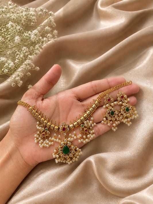 A close-up view of the Soudhamini Traditional Guttapusalu choker necklace and its matching drop earrings held gently in an open hand, showcasing the antique gold finish, emerald drop pendant, and delicate pearl fringe details against a shimmering silk background.