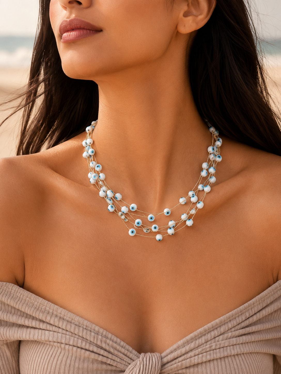 A flat-lay product photograph of a four-strand gold necklace adorned with small, white evil eye beads featuring blue and black centers. The necklace is elegantly arranged on a white marble slab, surrounded by soft champagne-colored satin fabric, a smooth beige stone, and a sprig of dried eucalyptus, emphasizing a serene and luxurious aesthetic.
