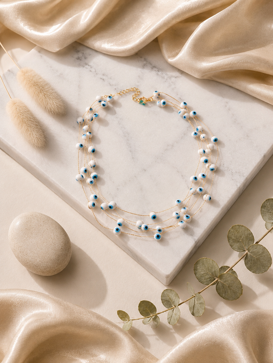 A flat-lay product photograph of a four-strand gold necklace adorned with small, white evil eye beads featuring blue and black centers. The necklace is elegantly arranged on a white marble slab, surrounded by soft champagne-colored satin fabric, a smooth beige stone, and a sprig of dried eucalyptus, emphasizing a serene and luxurious aesthetic.