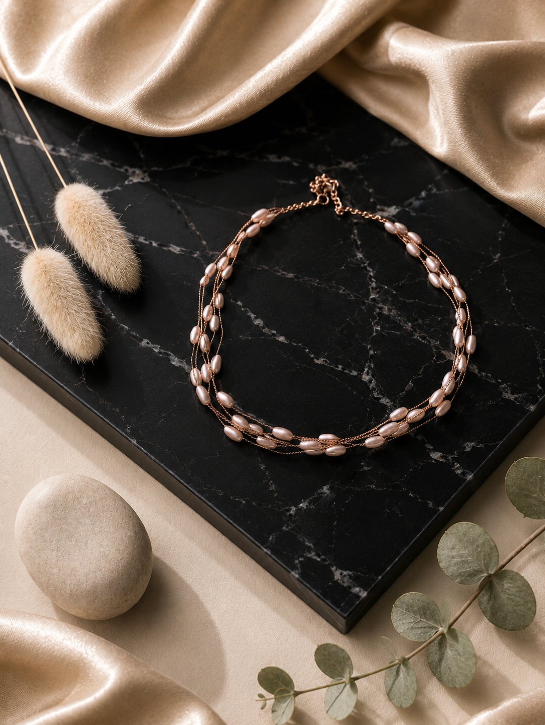 A high-angle product photograph from GS004_shell necklace_399.png showing a multi-strand gold necklace adorned with small, blush-pink oval shell beads, elegantly displayed on a black marbled slab flanked by shimmering champagne silk, dried bunny tails, and a smooth river stone.