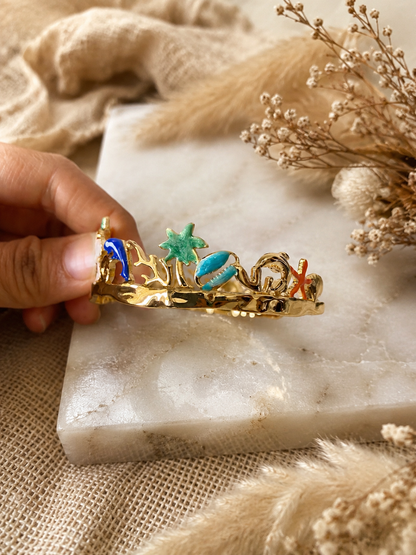 Gold bracelet with colorful coral and starfish designs held by a hand on a marble surface.