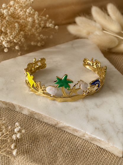 Gold-plated open cuff bracelet, featuring colorful enameled ocean charms like a green palm tree, starfish, white seahorse, and blue dolphin, beautifully displayed on a white marble slab surrounded by dried decorative flowers.