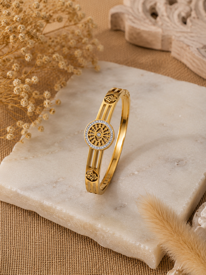 A flat-lay product photograph of a polished yellow-gold bangle bracelet resting on a textured white marble slab. The bangle features a central, circular sunwheel design with an evil eye center, bordered by small sparkling crystals, flanked by open-work Hamsa hand motifs on a triple-band frame. The background includes dried pampas grass and neutral woven textures. As seen in KD0023_sunwheel_a.png