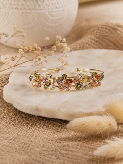 Gold open-cuff bangle bracelet featuring five floral charms inlaid with multi-colored gemstones in shades of green, orange, pink, and yellow, displayed on a white marble slab with a rustic burlap background.