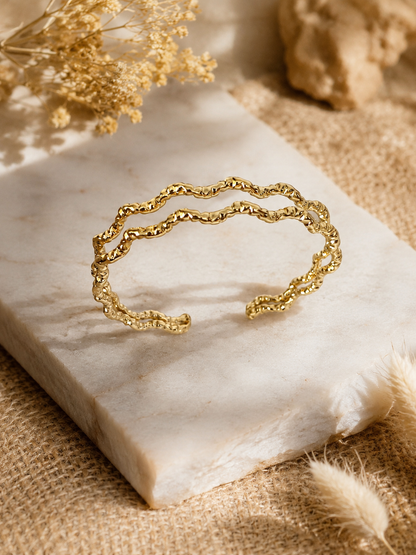 An open, double-banded gold cuff bracelet with a crinkled, wavy texture, displayed on a square white marble block surrounded by soft, neutral-toned dried florals and warm studio lighting.