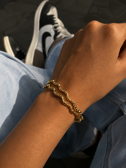 Gold bracelet on a wrist with a blurred background of a person wearing black and white sneakers.