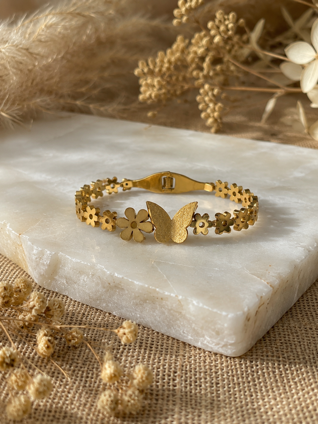 Gold chain bracelet featuring intricate cut-out flower links and a central matte-finish butterfly charm, displayed on a white marble slab with a rustic, neutral background