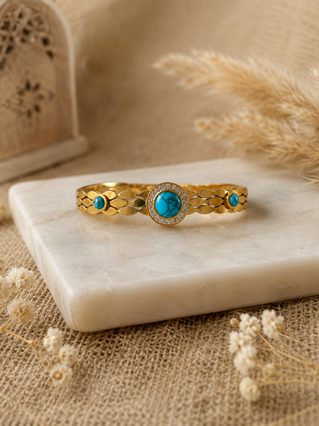A  bangle bracelet featuring a patterned scalloped band set with a central round turquoise stone surrounded by a crystal halo, accented by two smaller turquoise side stones, displayed on a white marble slab with dried florals.