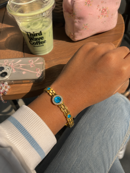 Hand wearing a gold bracelet with blue stones on a wooden table with a drink and phone case.