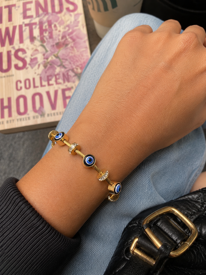Gold bracelet with blue beads on a wrist, with a book and coffee cup in the background.
