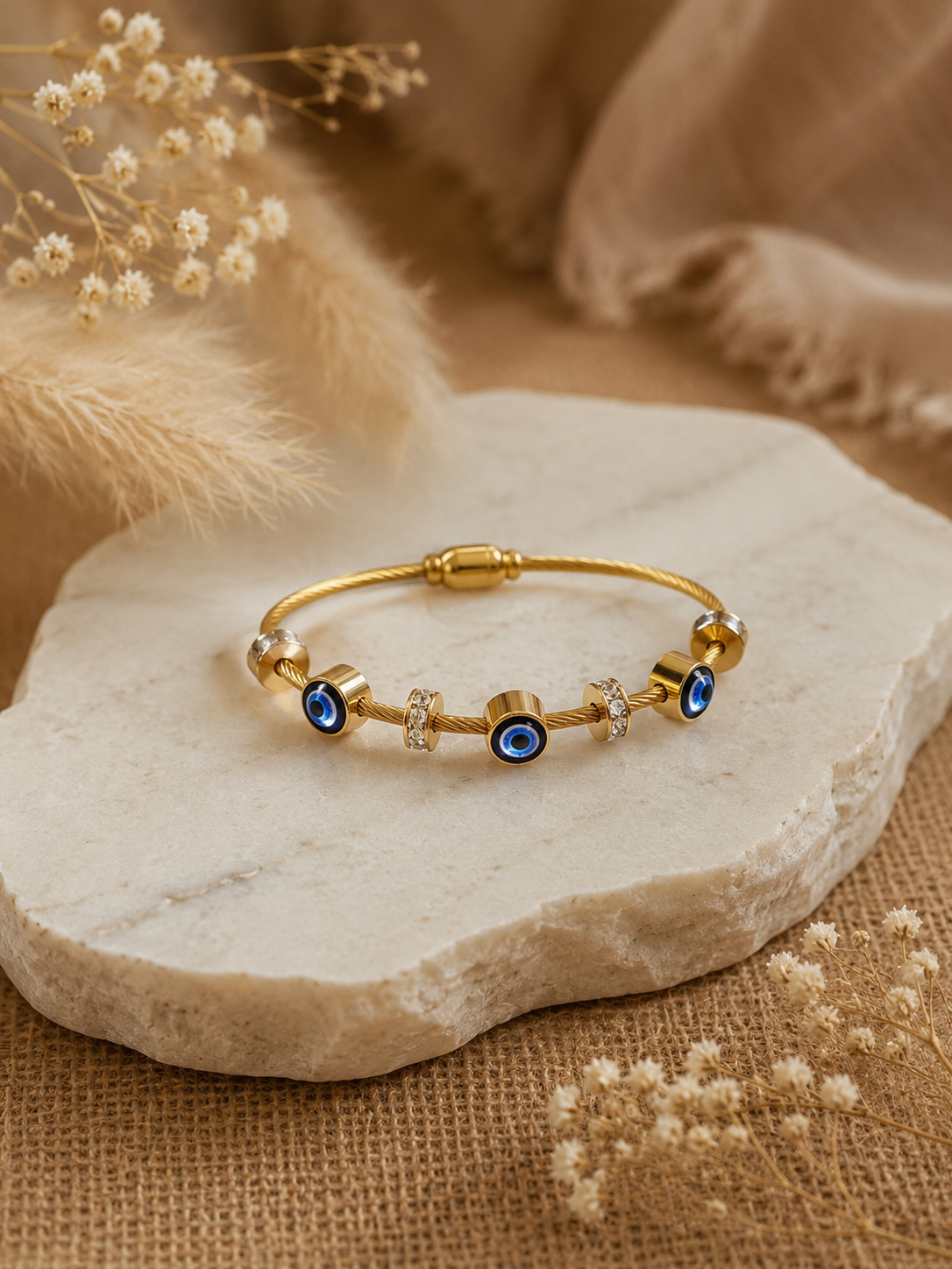 A gold twisted-cable bangle bracelet, adorned with three circular blue evil eye charms and crystal-studded spacer rings, resting elegantly on an organic-edged white marble slab surrounded by soft, beige dried florals.
