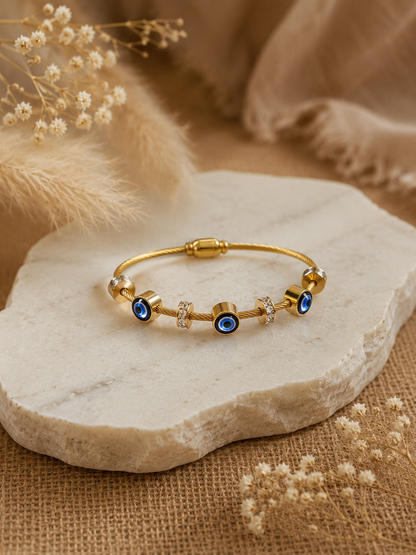 A gold twisted-cable bangle bracelet, adorned with three circular blue evil eye charms and crystal-studded spacer rings, resting elegantly on an organic-edged white marble slab surrounded by soft, beige dried florals.