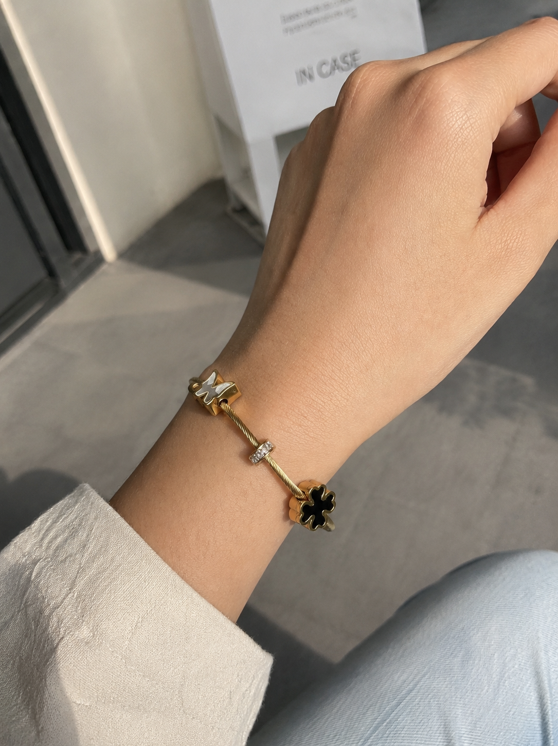 Hand wearing a gold bracelet with a black flower charm on a neutral background