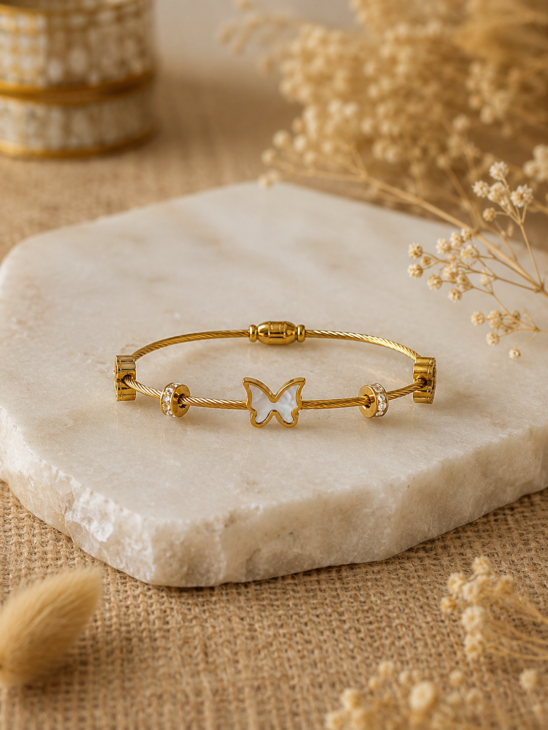 A gold twisted-cable bangle bracelet featuring a central gold butterfly charm with a white mother-of-pearl inlay, flanked by crystal-studded spacer beads, displayed elegantly on a raw-edged white marble slab surrounded by soft, neutral-colored dried flowers.
