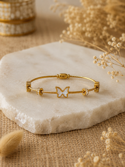 A gold twisted-cable bangle bracelet featuring a central gold butterfly charm with a white mother-of-pearl inlay, flanked by crystal-studded spacer beads, displayed elegantly on a raw-edged white marble slab surrounded by soft, neutral-colored dried flowers.
