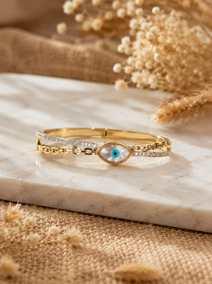 A gold double-layered bangle bracelet featuring a split band with one side pavé crystal-lined and the other styled like a gold link chain, joined by a central eye charm with a white mother-of-pearl inlay and blue center, resting on a white marble slab.