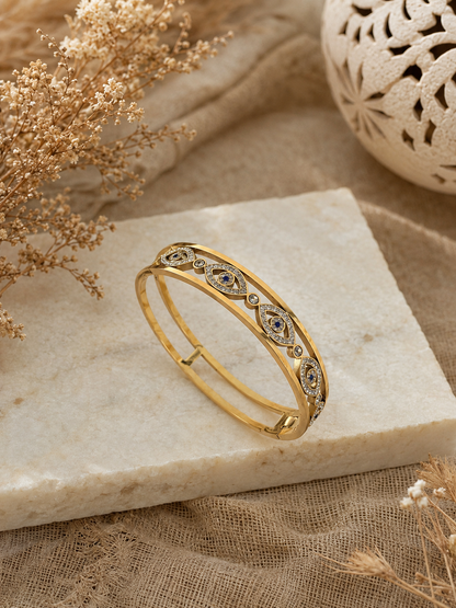 Antica Designs gold-toned evil eye bangle with micro-paved white crystals and blue center stones, worn on a wrist in a trendy, aesthetic café setting.