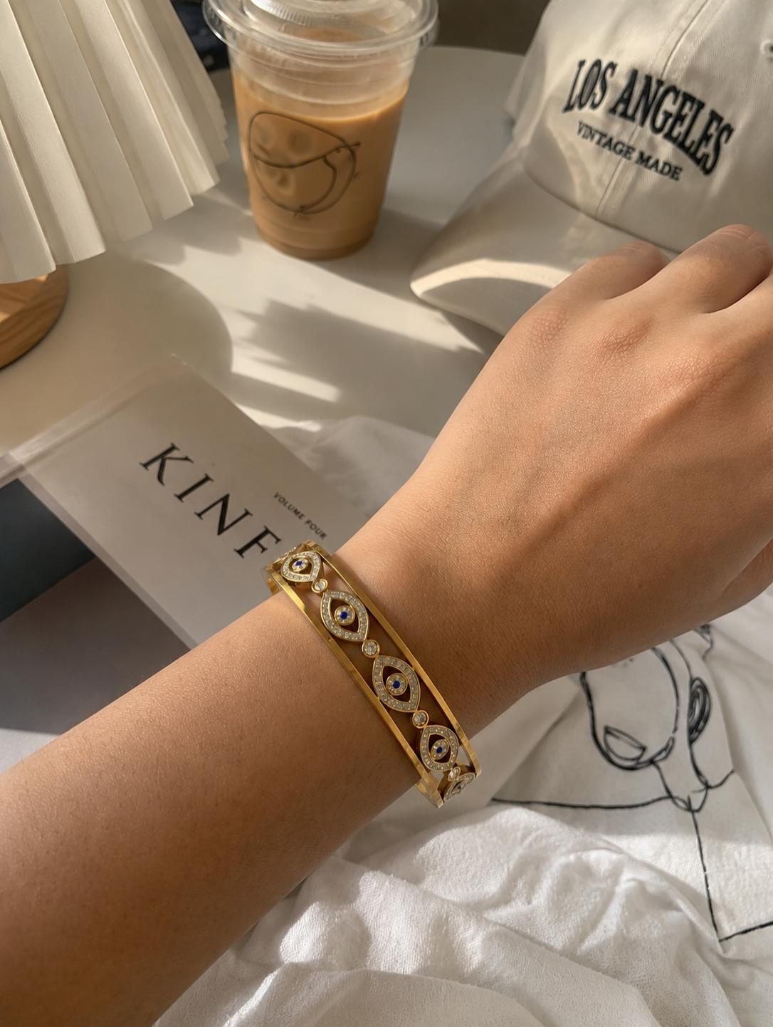 Antica Designs gold-toned evil eye bangle with micro-paved white crystals and blue center stones, worn on a wrist in a trendy, aesthetic café setting.