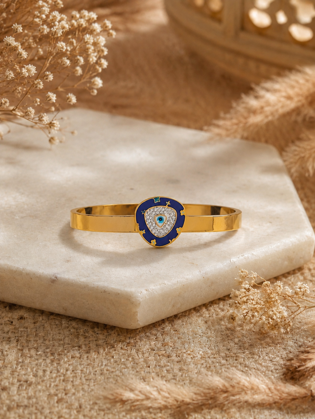 Antica Designs flat gold-toned cuff bracelet featuring a shield-shaped centerpiece with azure blue enamel, crystal pavé, and an evil eye motif displayed on a hexagonal stone plinth.